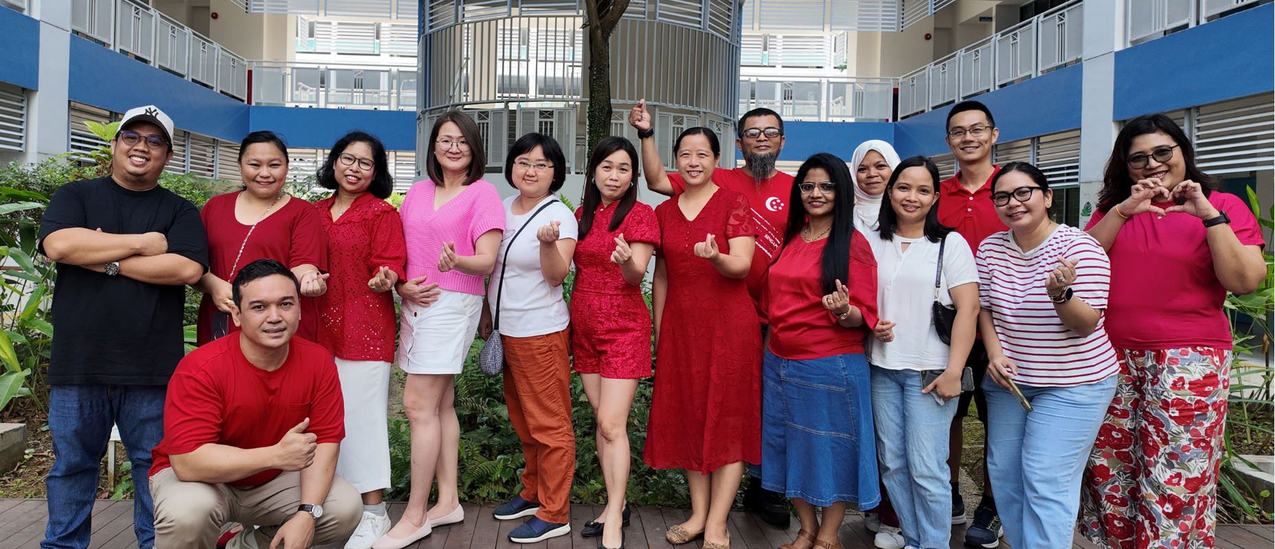 Group of 14 posing in a courtyard, many in red, making finger hearts; one person kneels in front.